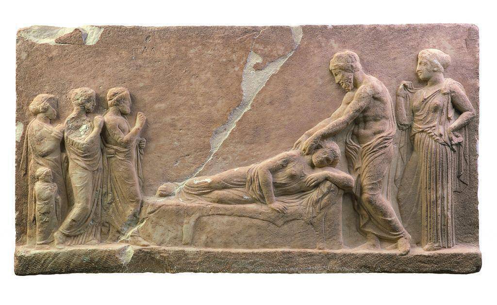 Μαρμάρινο αναθηματικό ανάγλυφο: Υπό το βλέμμα της Υγιείας, ο Ασκληπιός θεραπεύει την ξαπλωμένη ασθενή κατά τη διάρκεια της εγκοίμησης στο Ασκληπιείο. περ. 400 π.Χ., Αρχαιολογικό Μουσείο Πειραιά. © ΥΠΠΟΑ – ΚΣΤ΄ΕΠΚΑ (φωτογρ. αρχείο) 